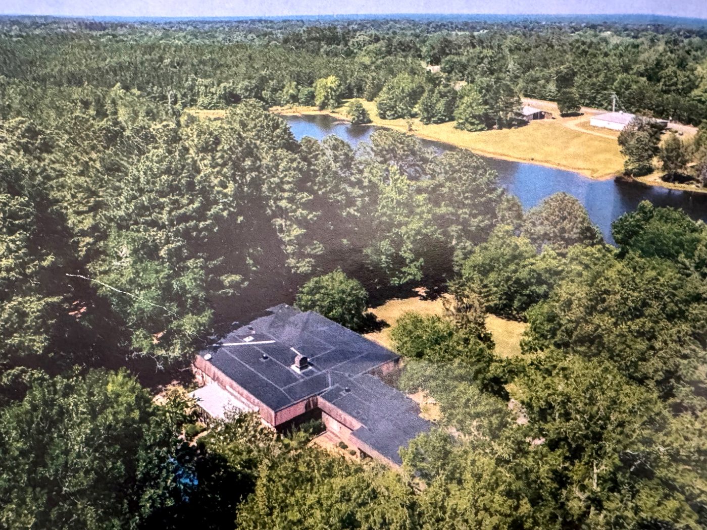 Aerial view of the long brick house among mature trees, with open lawn beside it and a lake beyond