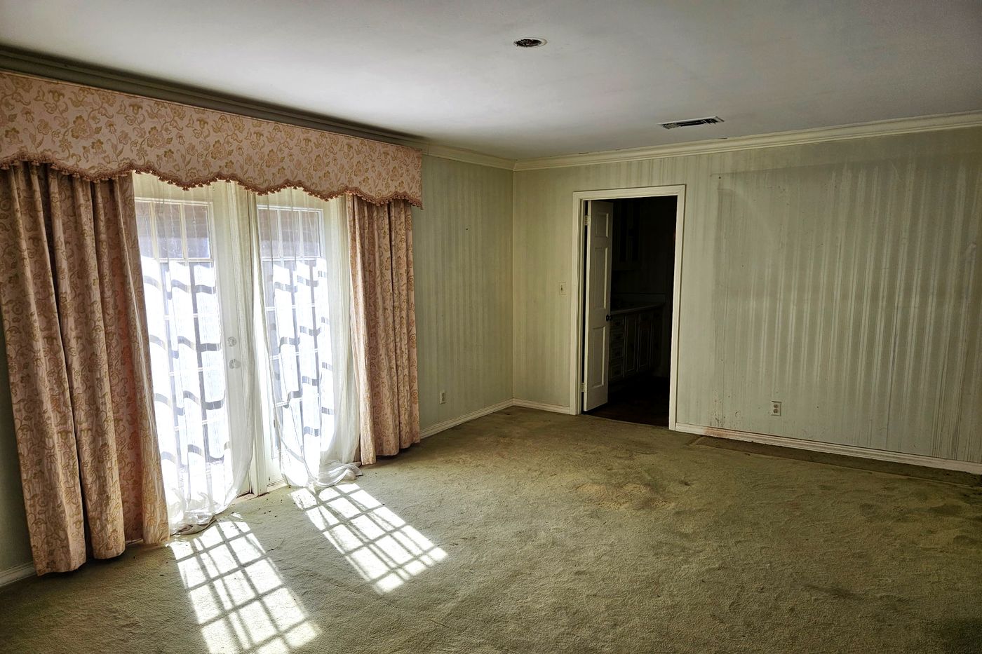 Carpeted bedroom with striped wallpaper, crown molding, and French doors hung with floral draperies, sunlight falling across the floor