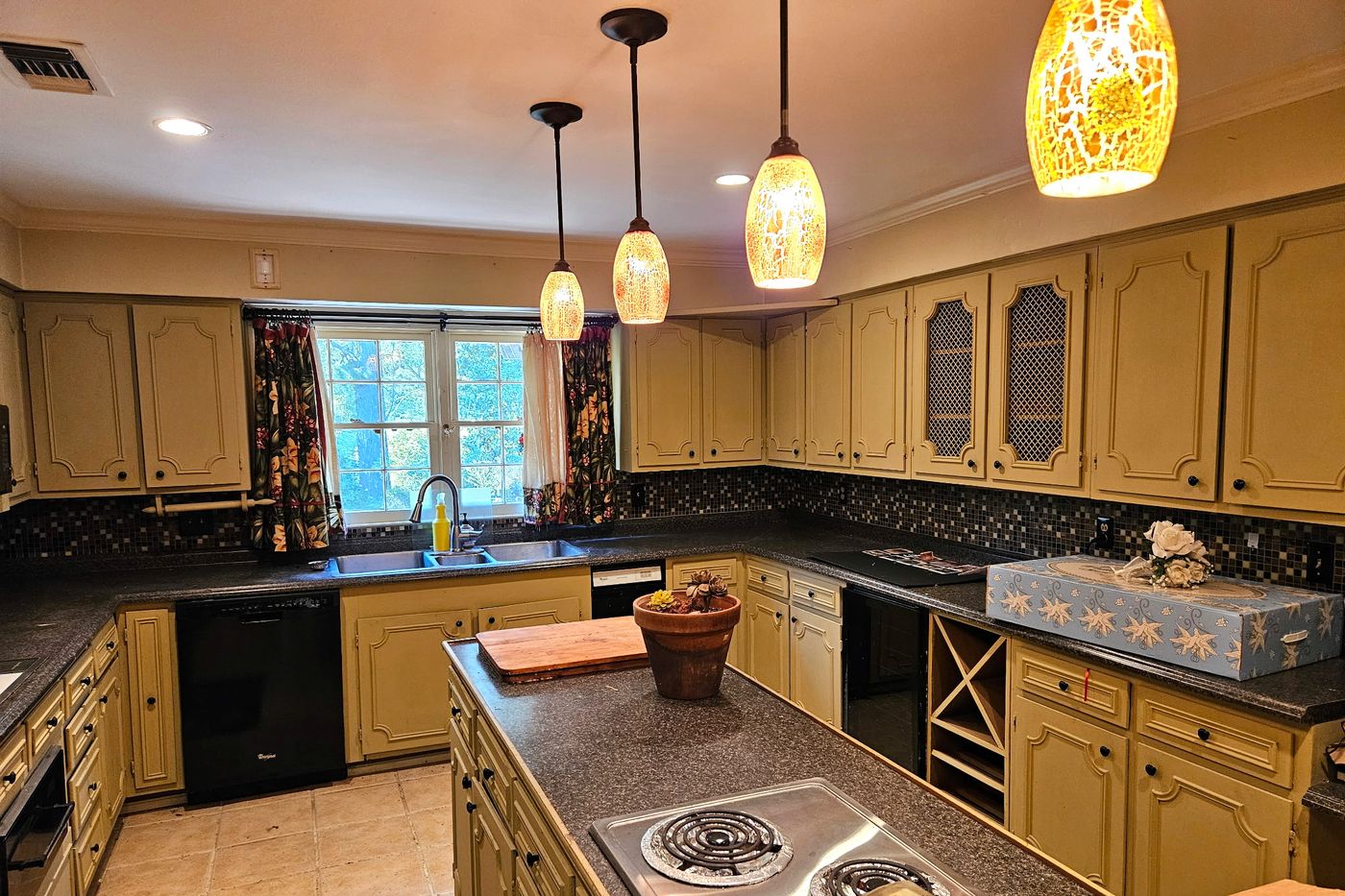 Kitchen with painted raised-panel cabinetry, dark countertops, a mosaic tile backsplash, a window over the sink, pendant lights, and a center island with an electric cooktop