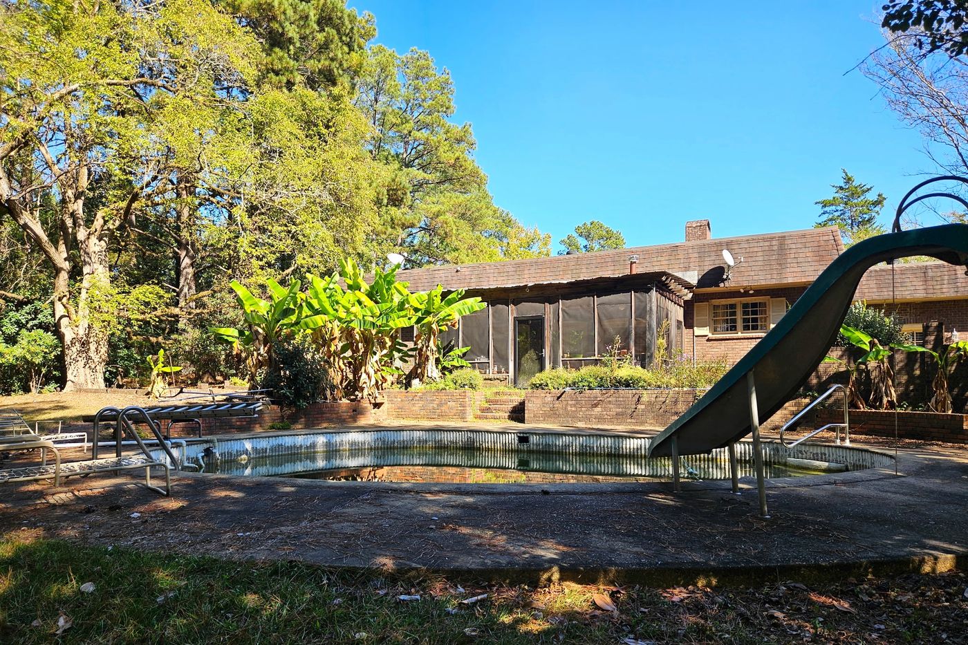 In-ground pool with a slide and low green water, in need of restoration, in front of the screened porch and brick rear of the house